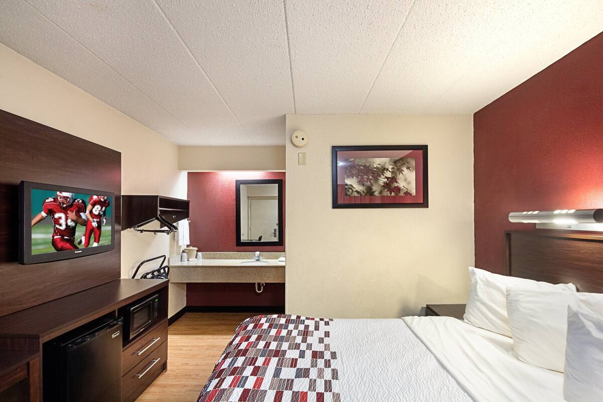 A hotel room with a bed, a TV mounted on the wall, a desk with a computer, a mirror, and a picture on the wall.