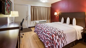 Ein Hotelzimmer mit Bett, Schreibtisch, Stuhl, Lampe, Wandfernseher und einem Fenster mit Vorhängen.