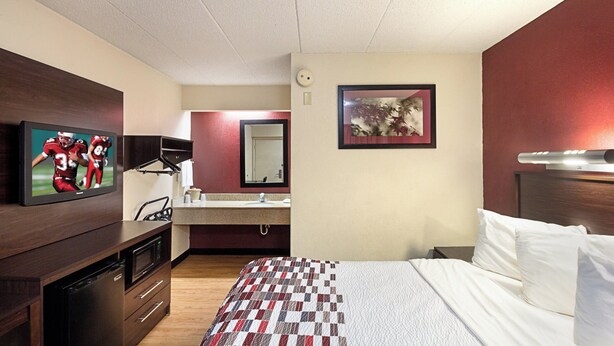 A hotel room with a bed, a TV mounted on the wall, a bathroom with a mirror, and a picture on the wall. - Red Roof Inn Cincinnati East - Beechmont (Cincinnati)