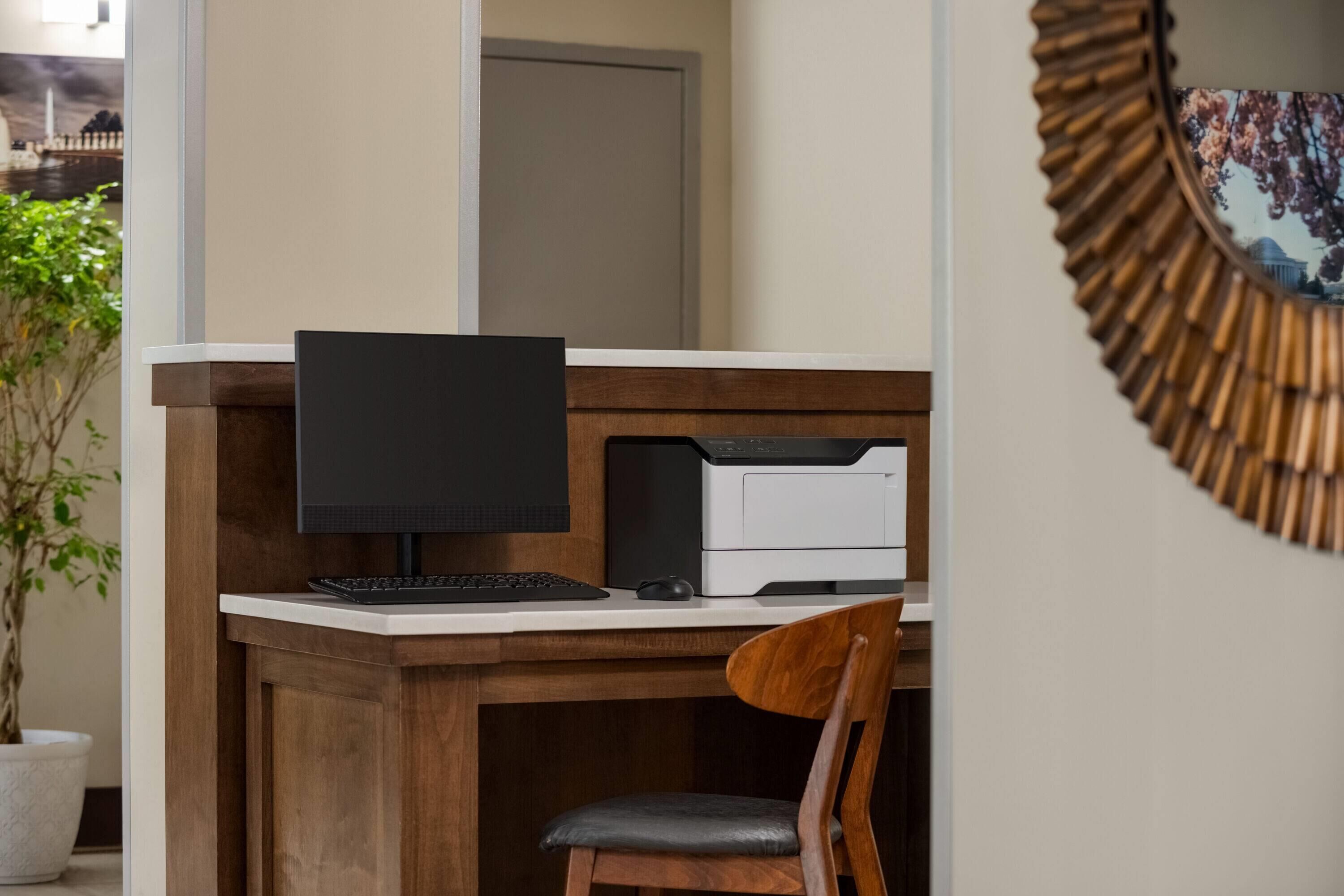 A wooden desk with a computer monitor, keyboard, and printer. A chair is positioned in front of the desk.