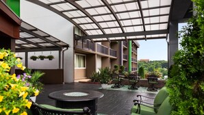 Un espacio al aire libre techado con una mesa redonda y sillas verdes, rodeado de plantas en macetas y una vista de un edificio al fondo.