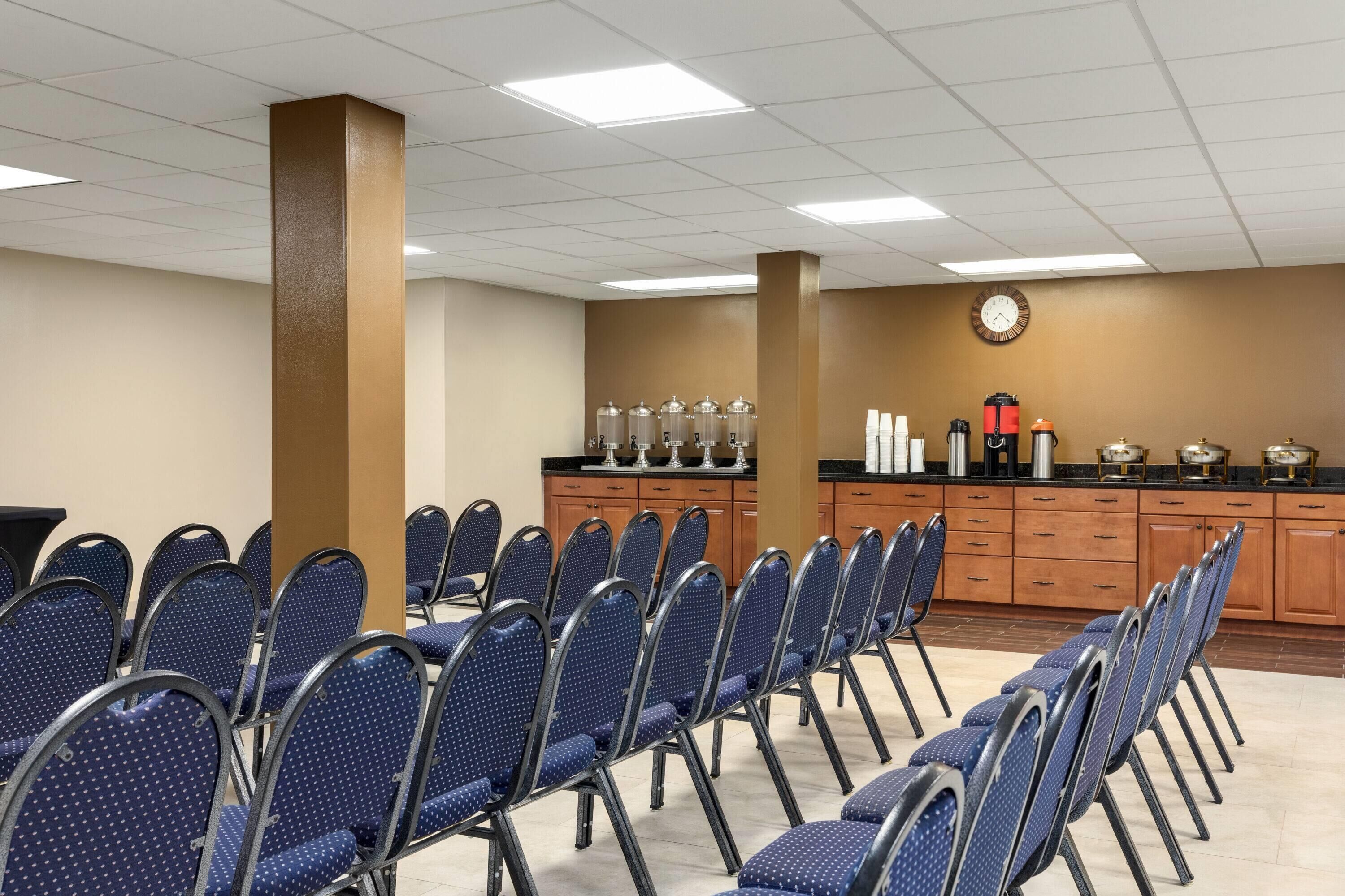 Une salle de conférence avec des rangées de chaises bleues, un coin cuisine équipé d’appareils électroménagers et une horloge au mur.