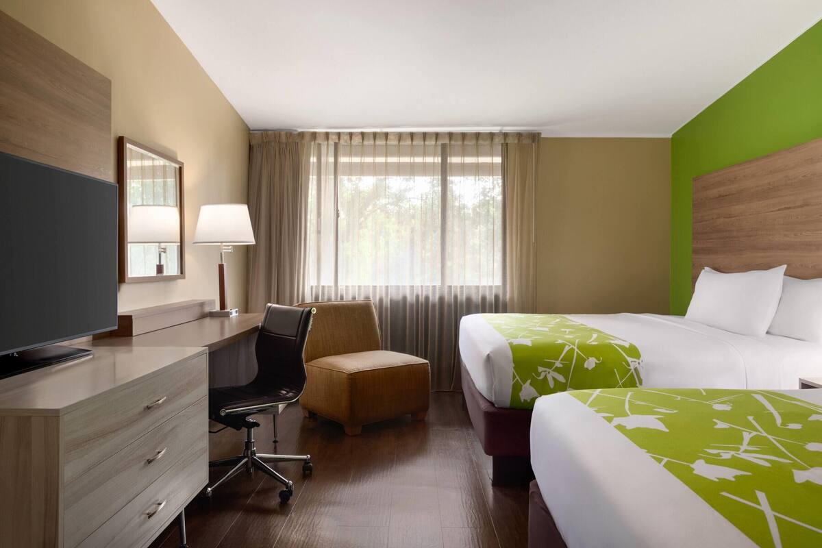 Una camera d'albergo con scrivania, sedia, due letti, televisore e finestra con tende.