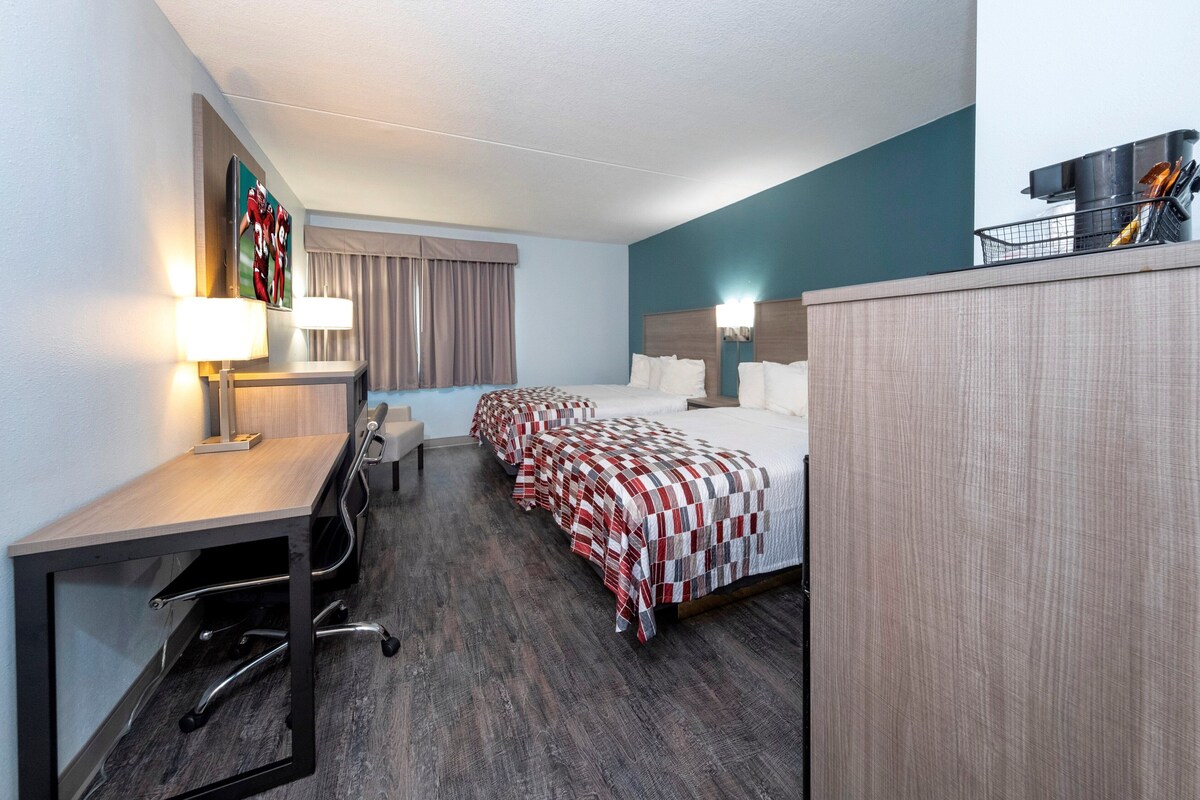 Une chambre d’hôtel équipée d’un bureau, de deux lits, d’une tête de lit, d’une fenêtre avec des rideaux et d’une lampe fixée au mur.
