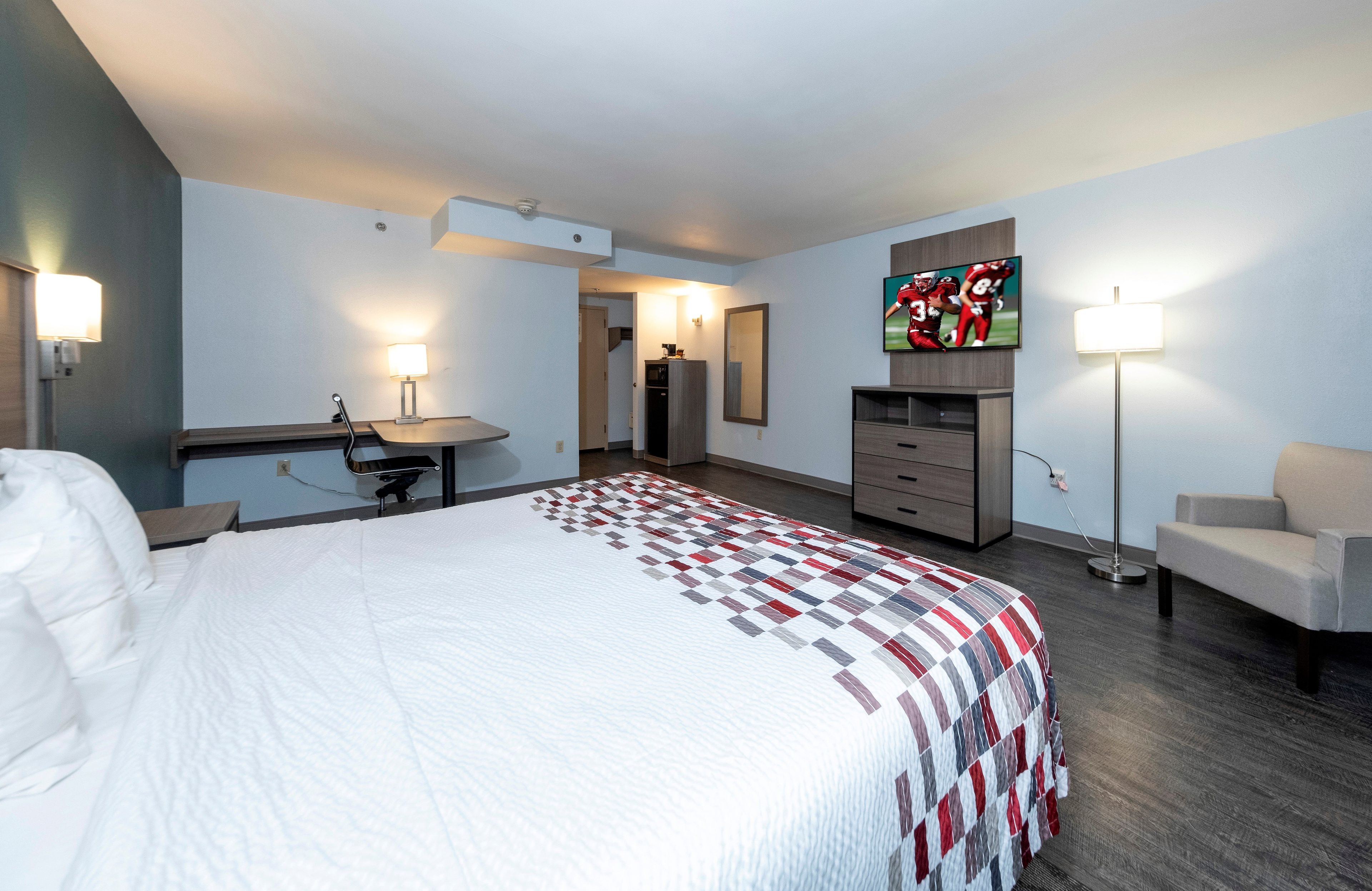 A hotel room with a bed, a desk, a chair, a TV, and a lamp.