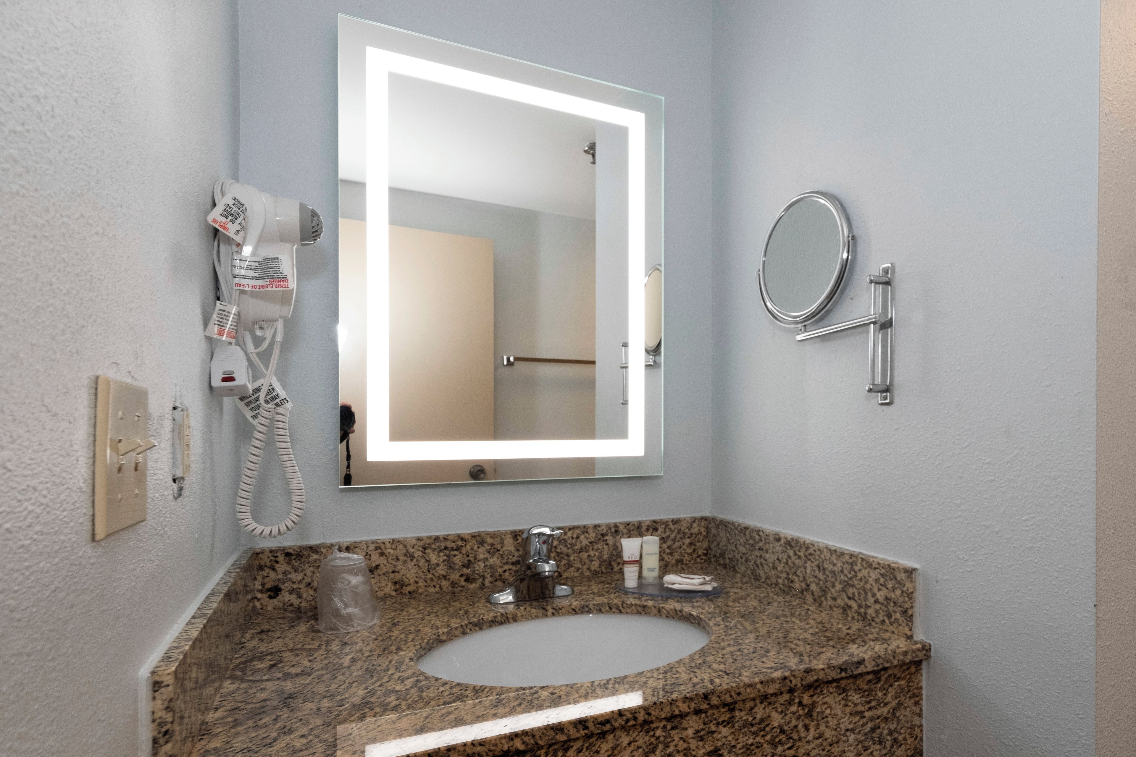 A bathroom with a granite countertop, a sink, a mirror, a handheld hair dryer, and a wall-mounted telephone.