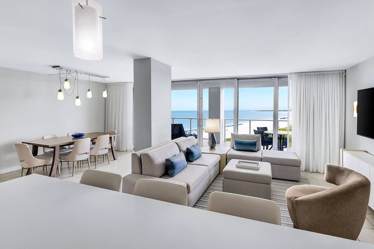 A modern hotel room with a dining area, a sofa set, a flat-screen TV, and a view of the sea.