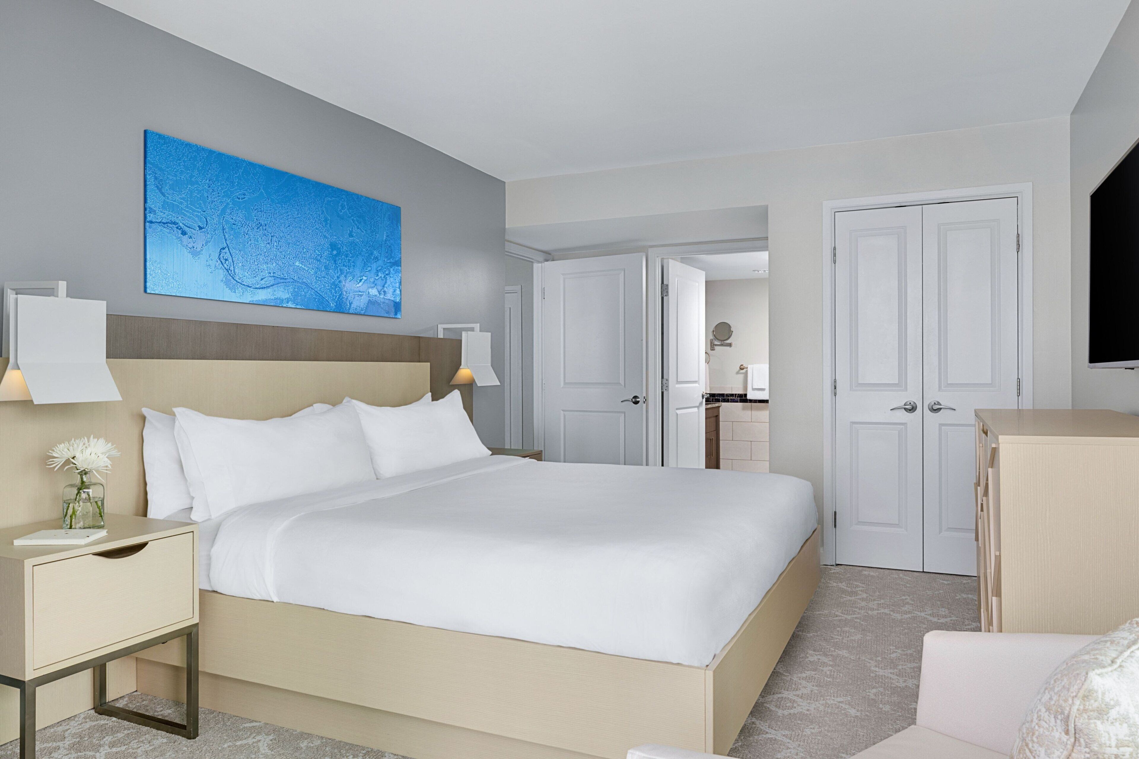 A hotel room with a large bed, a bedside table with a lamp, a small table with a vase, and a television on the wall.
