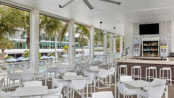 Uma área de estar moderna ao ar livre com mesas e cadeiras brancas, piscina ao fundo e um bar com painel de pedra.