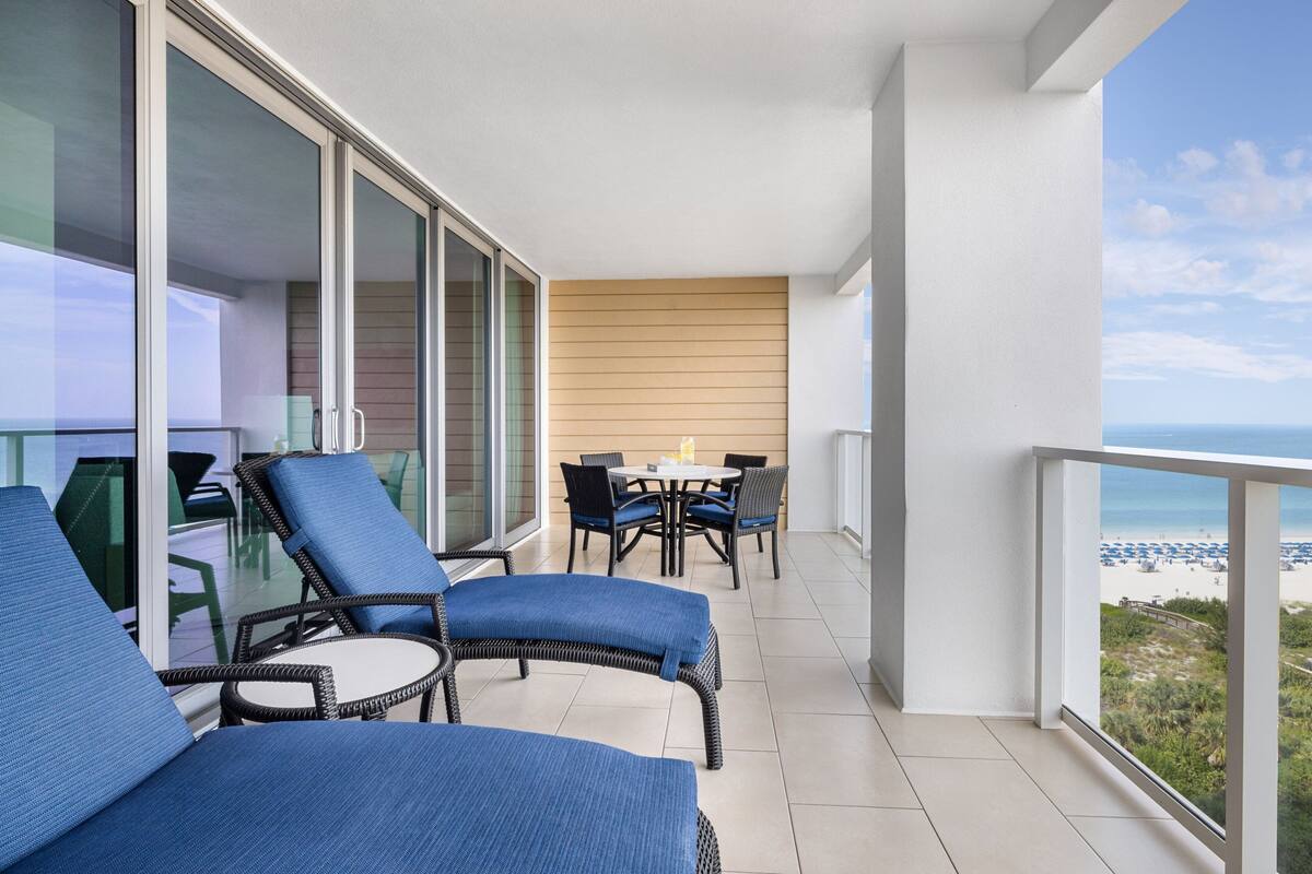 A balcony with wicker chairs and a table, overlooking a beach.