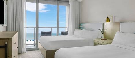 A hotel room with two beds, a TV, a desk, and a view of the ocean.