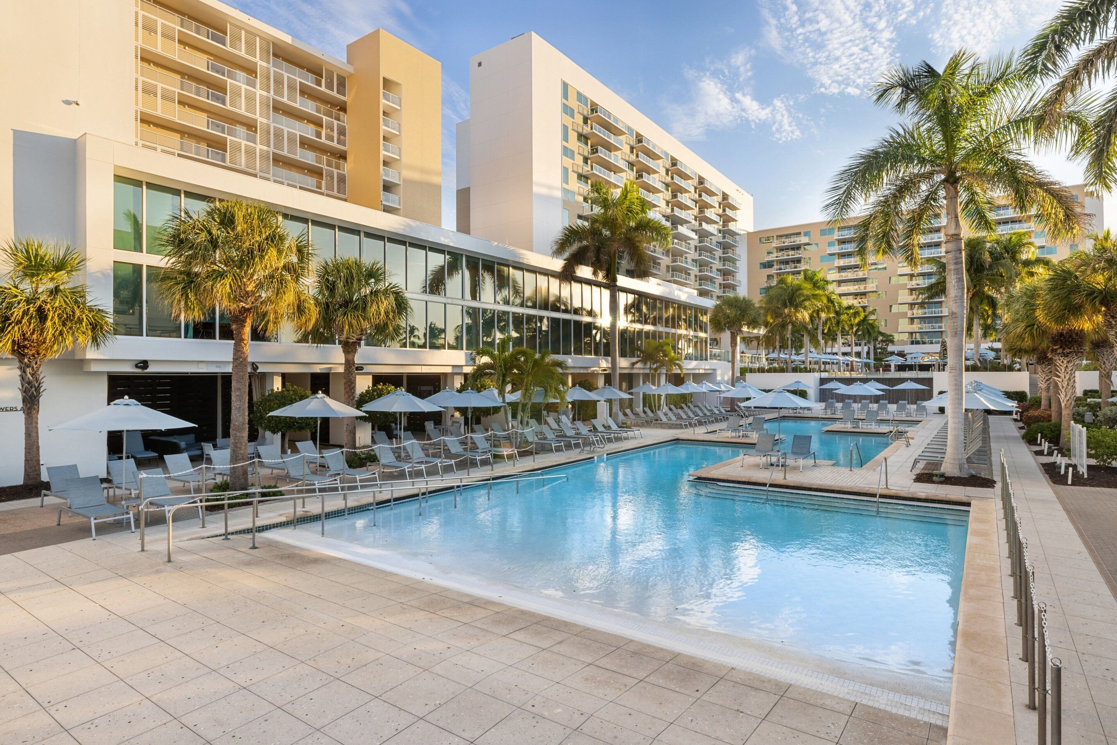 Un espace piscine d’hôtel avec des chaises longues et des parasols, entouré de palmiers et de bâtiments modernes.
