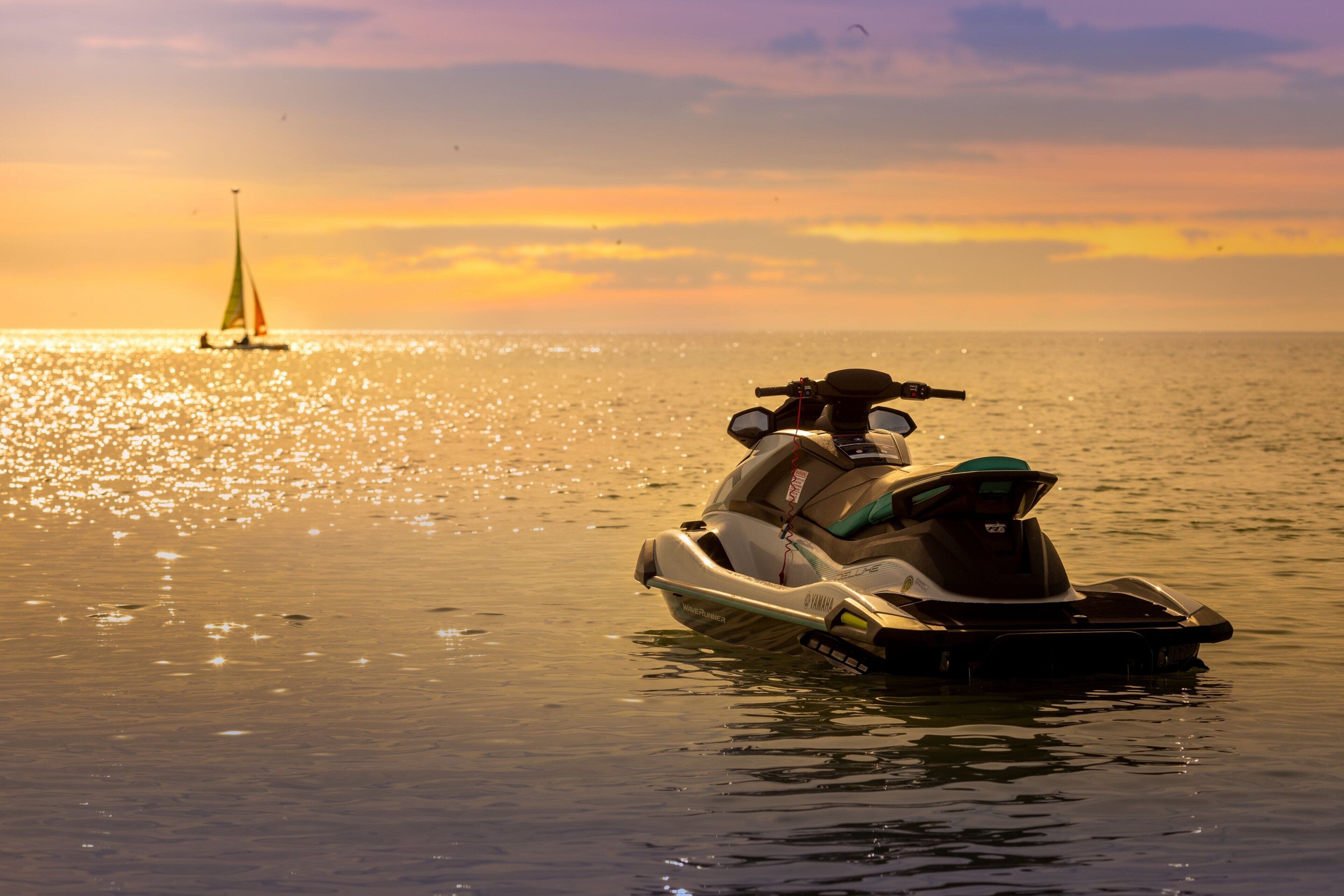A jet ski on calm water with a sailboat in the distance during sunset.