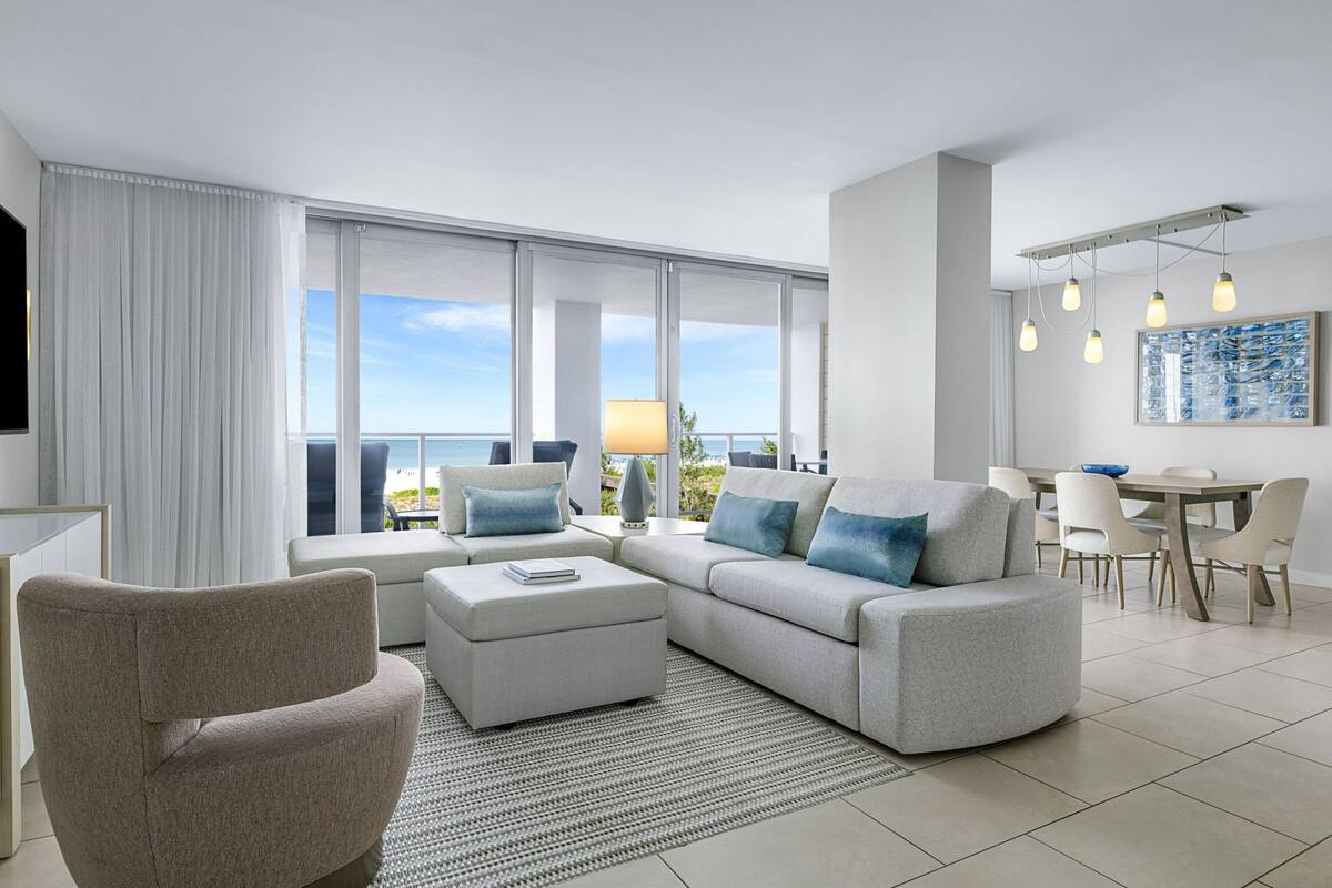 A modern living room with a sofa, ottoman, and armchair, a dining area with a table and chairs, and large windows offering a view of the ocean.
