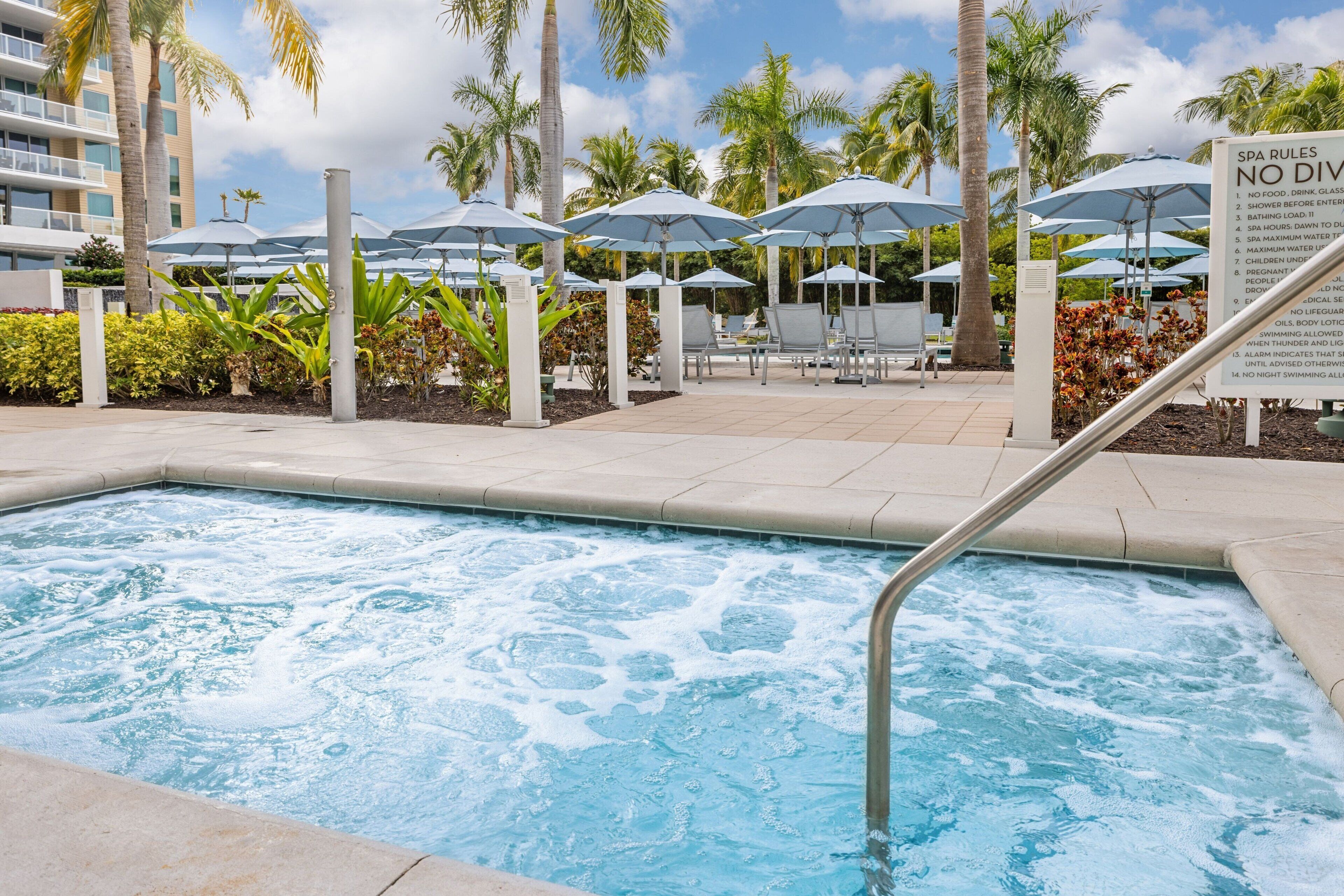Une piscine avec un jacuzzi, entourée de parasols et de chaises longues, et un panneau indiquant les règles du spa.