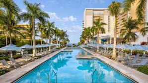 Ein Swimmingpool mit Liegestühlen und Sonnenschirmen, umgeben von Palmen und einem Hotelgebäude.