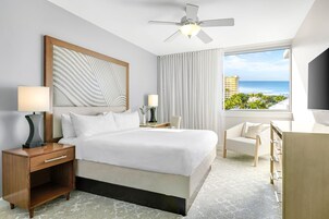Quarto de hotel com uma cama grande, uma escrivaninha, uma cadeira, uma mesa de cabeceira, um ventilador de teto e vista para o mar.