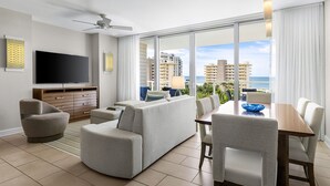 Ein modernes Wohnzimmer mit Fernseher, grauen Sofas, einem Holzesstisch und einem großen Fenster mit Blick auf Stadt und Meer.