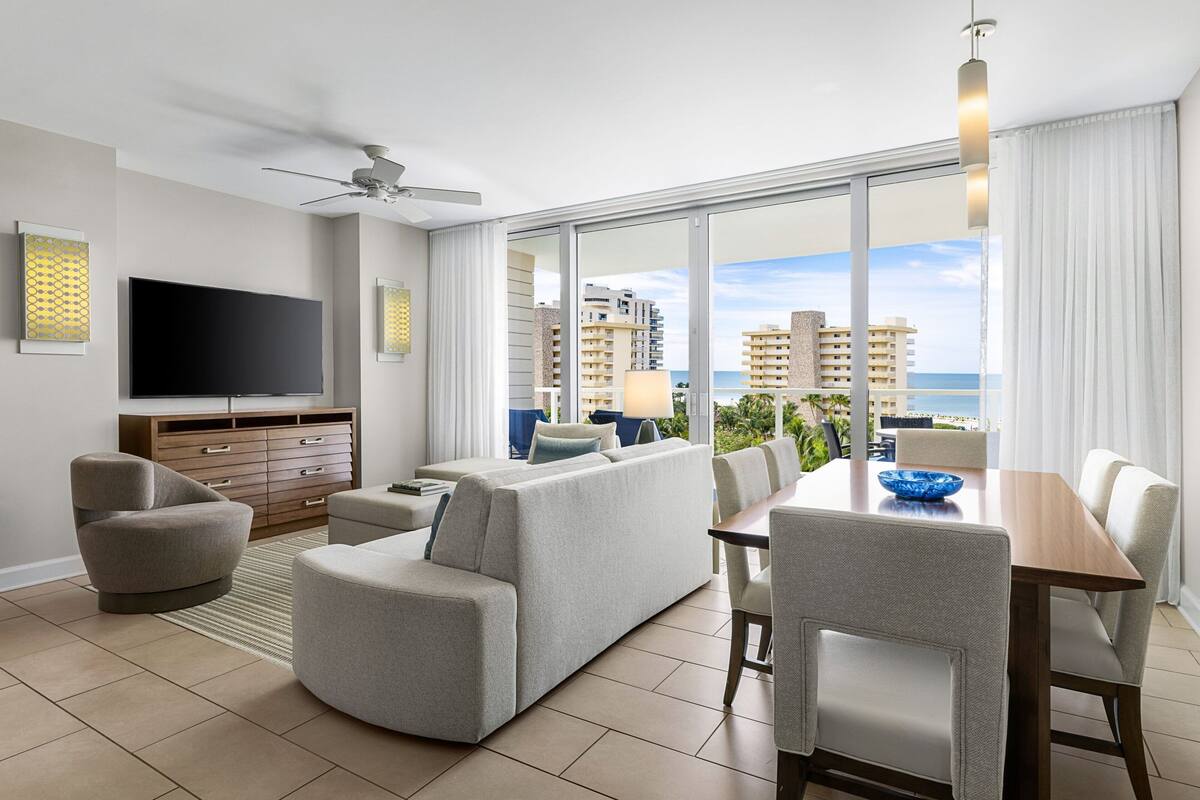 A modern living room with a flat-screen TV, grey sofas, a wooden dining table, and a large window offering a view of the city and ocean.