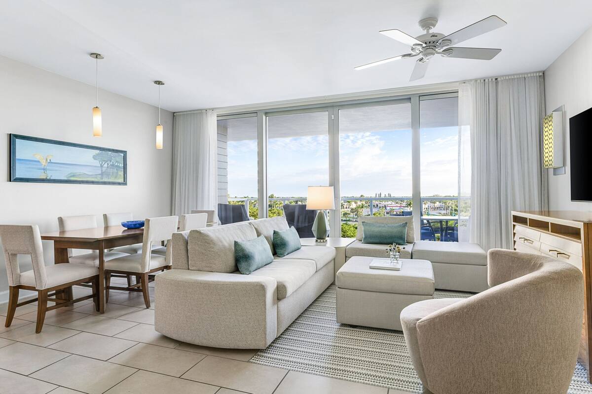 A modern hotel room with a large window, a dining area, a sofa, a TV, and a ceiling fan.