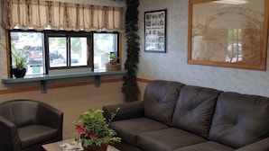 A living room with a brown leather couch, a wooden coffee table with magazines, a potted plant, and a window with a view of parked cars.