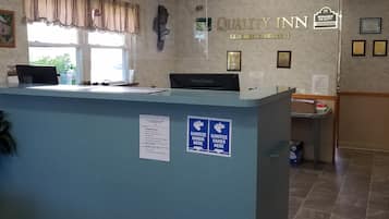 A reception desk with a sign reading "QUALITY INN BY CHOICE HOTELS" and a "MANAGER ON DUTY" sign. There are two hand sanitizer signs on the wall, a ViewSonic monitor, and a framed certificate.