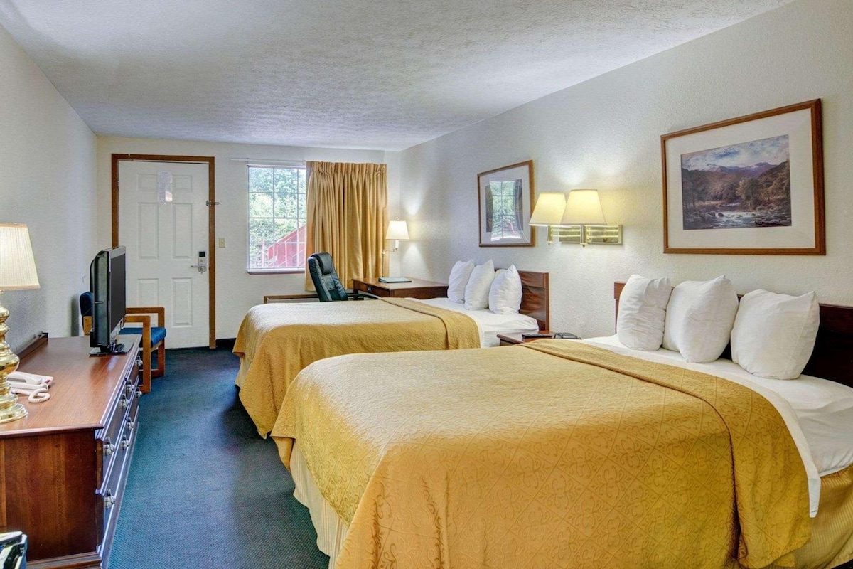 A hotel room with two beds, a desk with a lamp, a chair, a television, and a window with curtains.