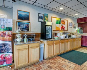 A breakfast buffet area with a coffee machine, fruit display, and a refrigerator.