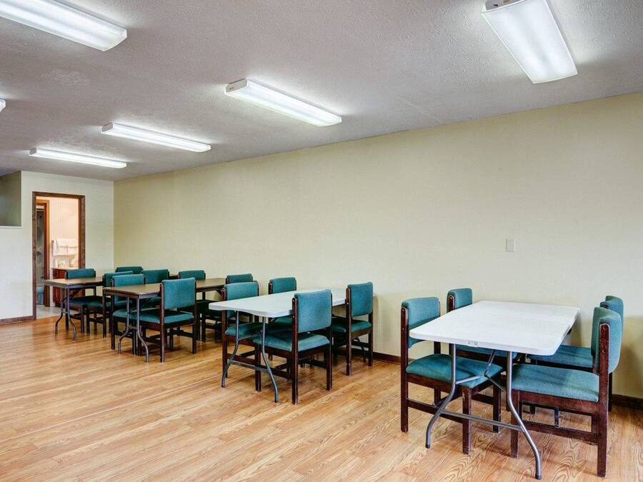 A room with wooden flooring, teal upholstered chairs, and white tables arranged in a U-shape.
