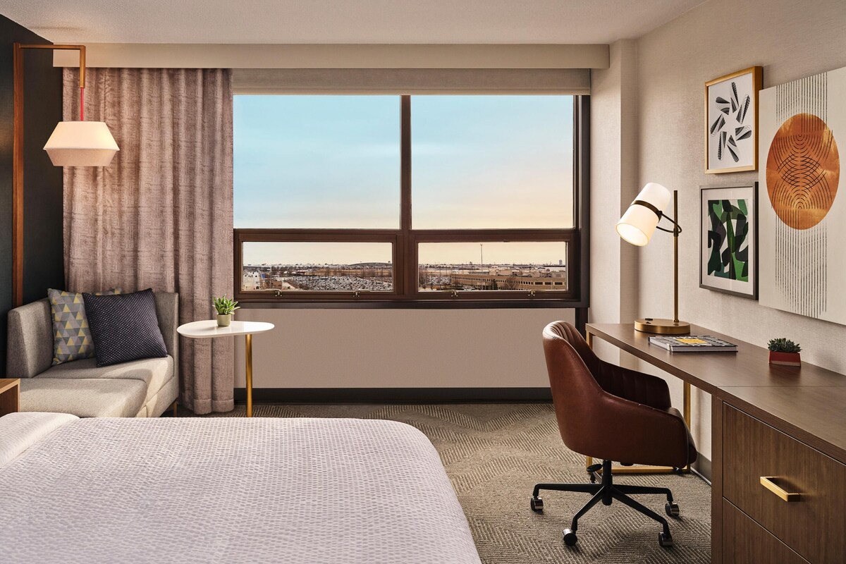 Ett hotellrum med en säng, ett skrivbord, en stol, ett litet bord, en lampa, en växt och ett stort fönster med utsikt över staden.