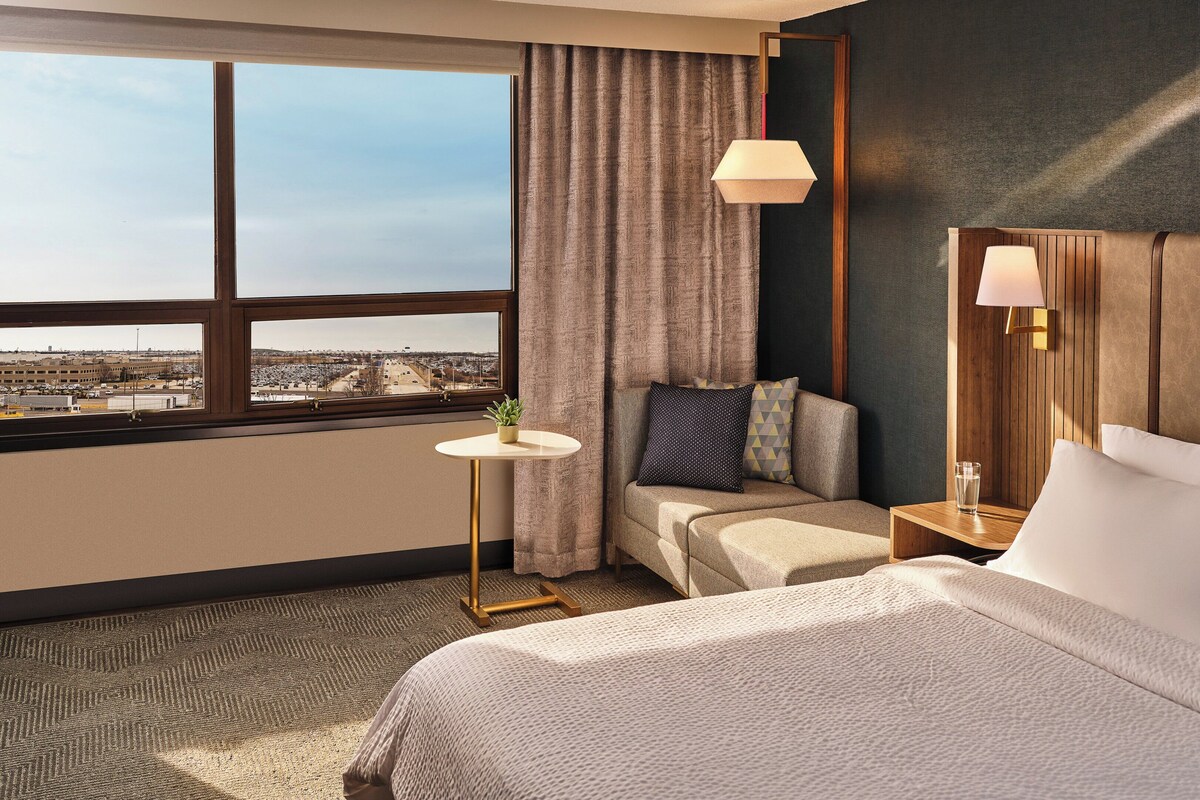 Ein Hotelzimmer mit großem Fenster, einer Couch, einem Nachttisch mit Lampe und einem Bett mit Kissen.