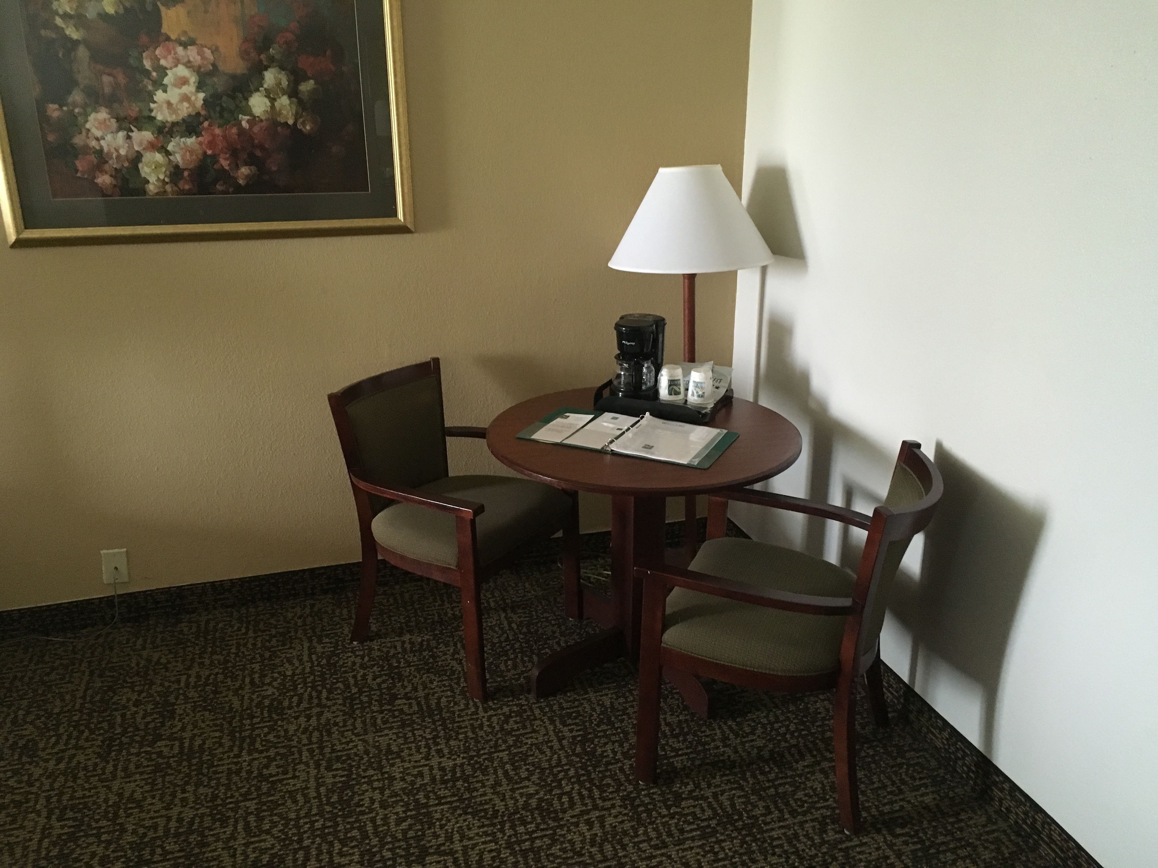 Habitación de hotel con una mesa redonda, dos sillas, una lámpara, cafetera y un cuadro floral enmarcado en la pared.