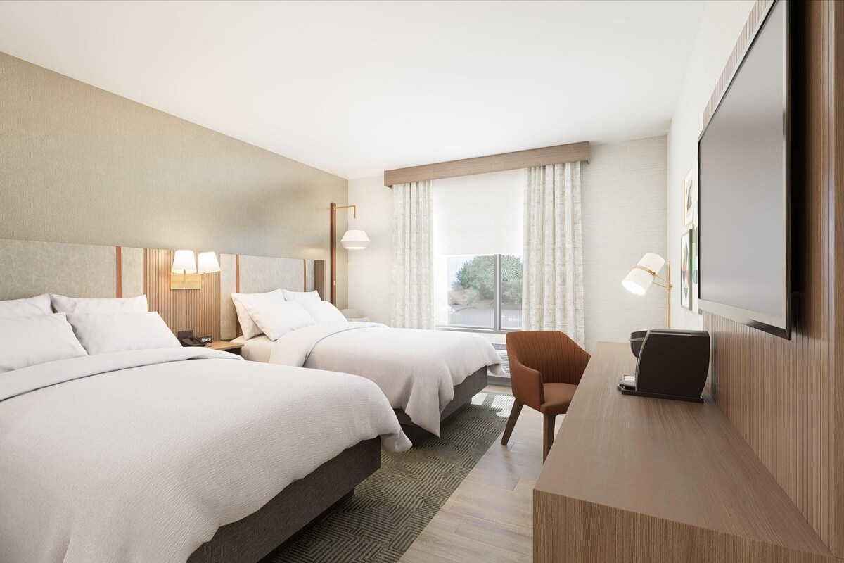 Ett hotellrum med två sängar, ett skrivbord, en stol, en TV och ett fönster med gardiner.