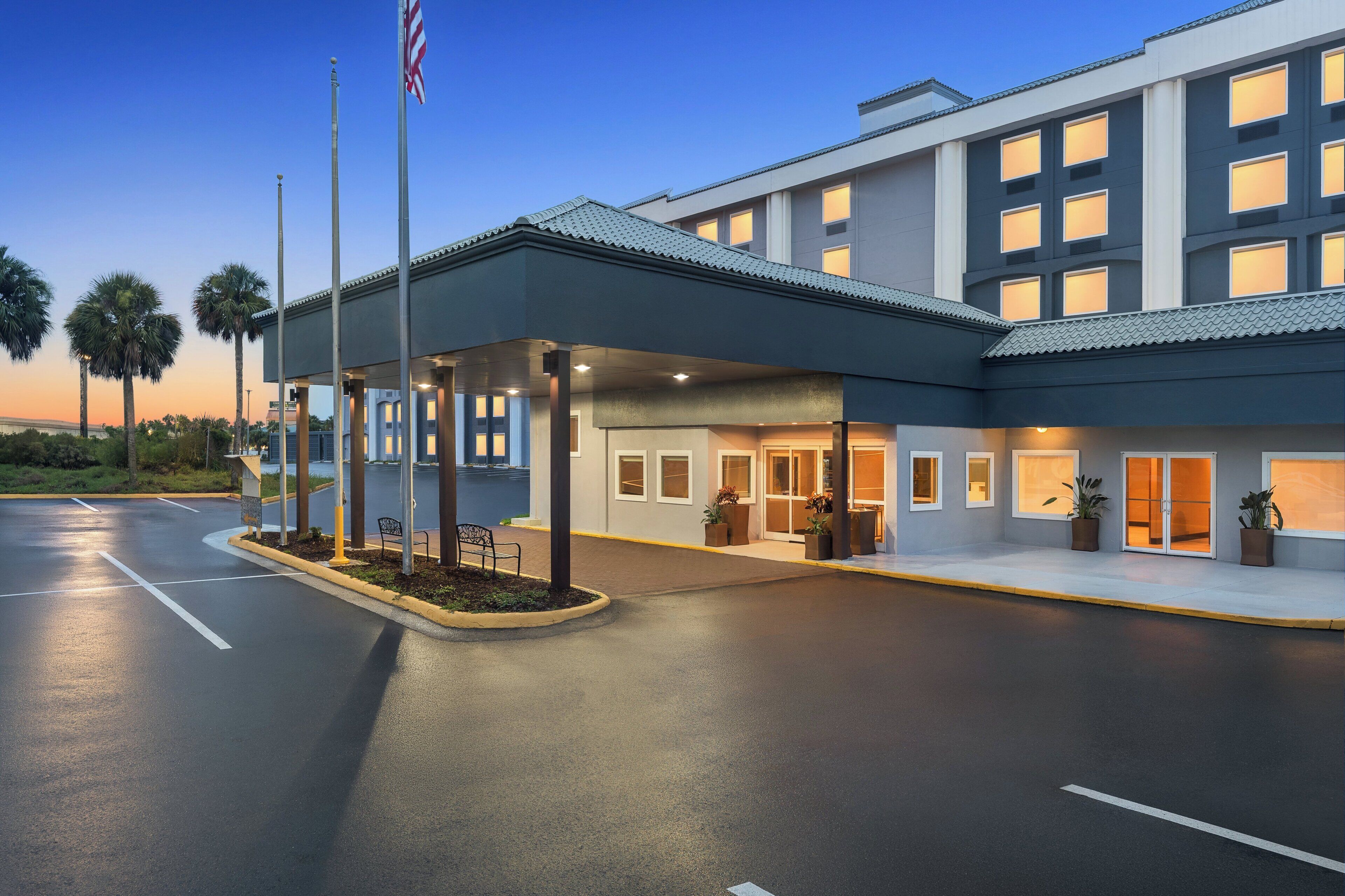 A modern hotel entrance with a covered waiting area, palm trees, and an American flag.