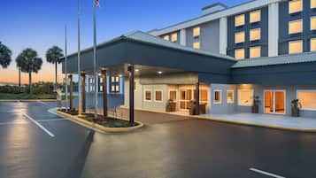 A modern hotel entrance with a covered waiting area, palm trees, and an American flag.