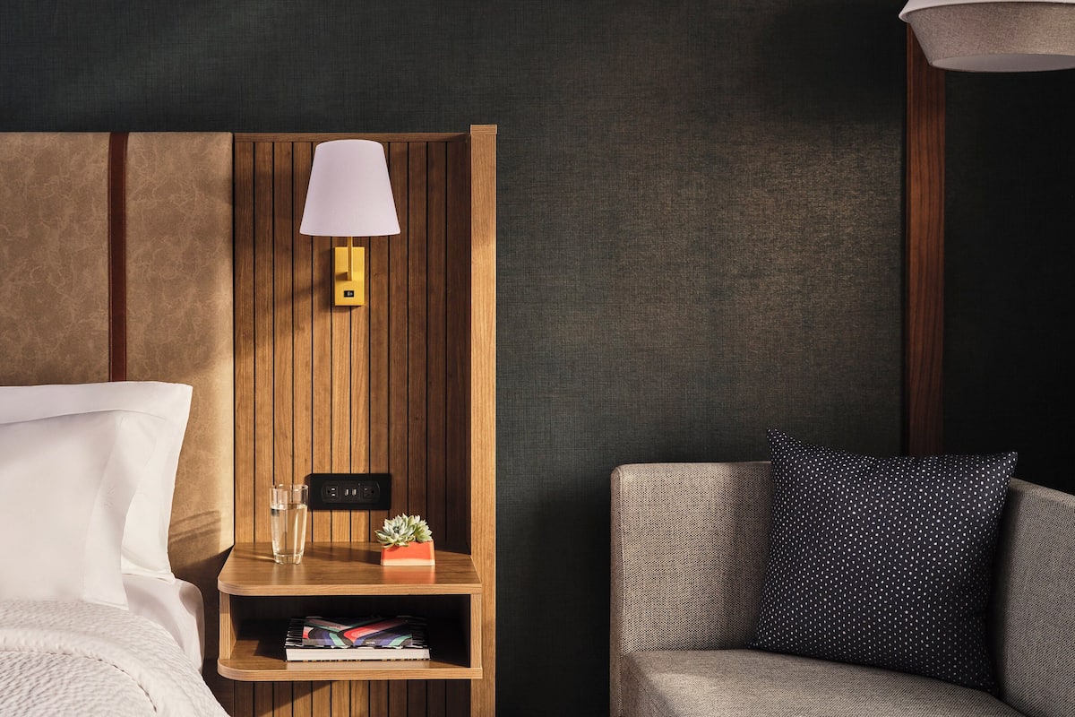A hotel room with a wooden headboard, a bedside table with a lamp, a small plant, and a bookshelf.