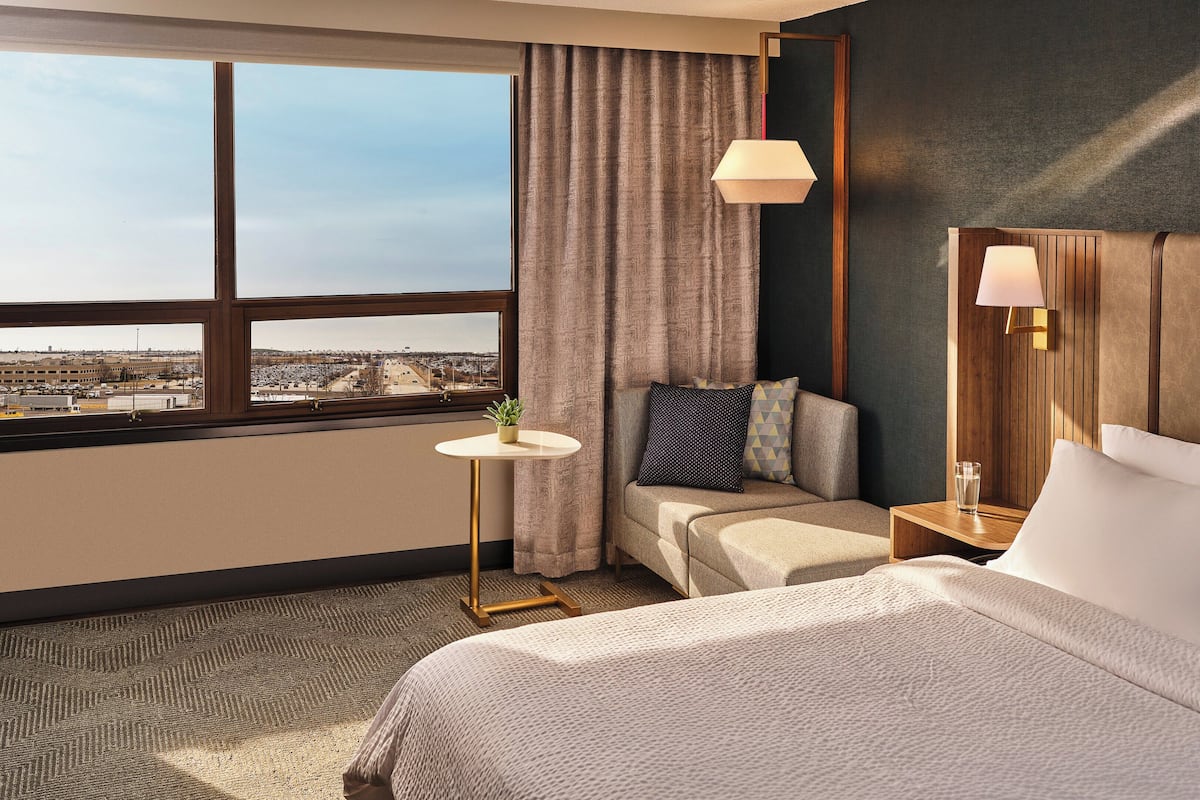 A hotel room with a large window, a sofa, a bedside table with a lamp, and a bed with pillows.