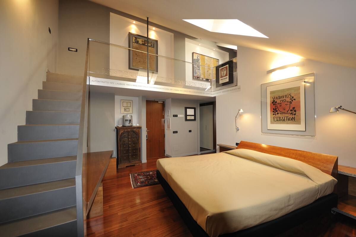A bedroom with a large bed, a staircase, and a loft area with artwork.