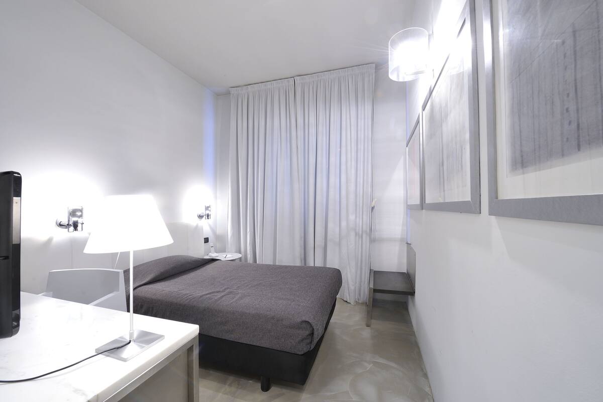 A modern hotel room with a bed, a desk with a lamp, a flat-screen TV, and a large window with curtains.