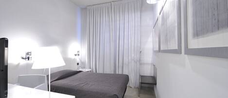 A modern hotel room with a bed, a desk with a lamp, a flat-screen TV, and a large window with curtains.