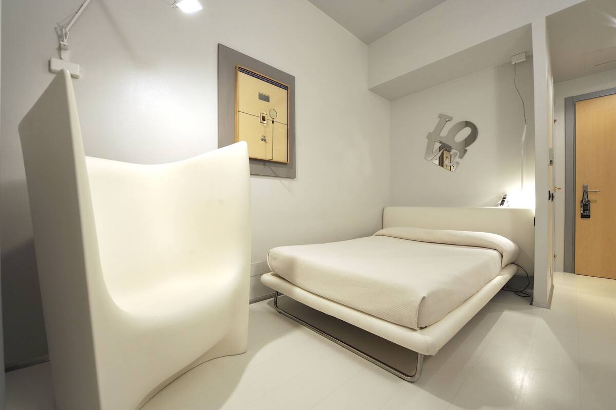 A modern bedroom with a bed, a chair, and a wall-mounted safe.
