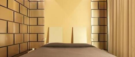 A modern bedroom with a bed centered on a platform, flanked by brick-patterned walls and a large window with curtains.