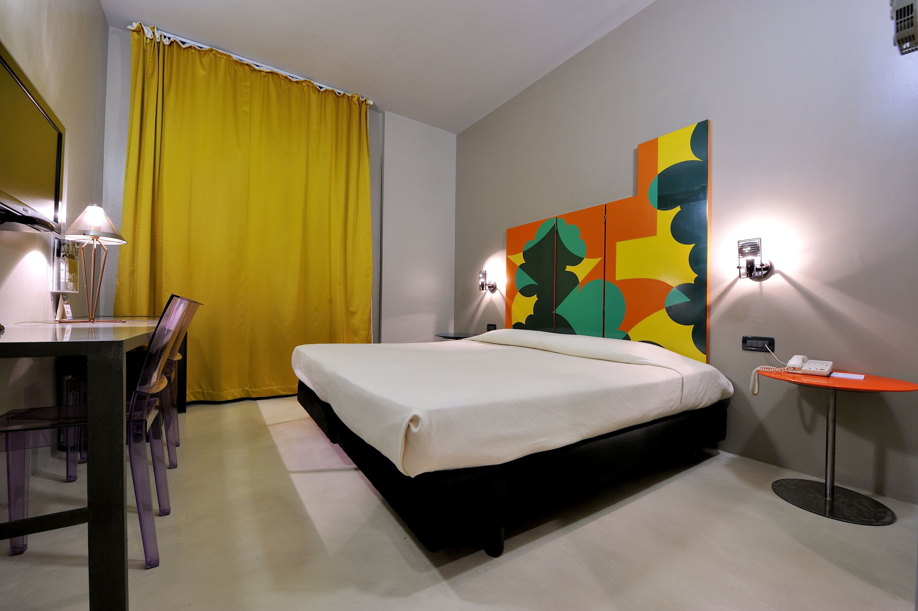 Une chambre d’hôtel avec un lit, un bureau doté d’une lampe, une chaise, une petite table avec un téléphone et une œuvre d’art colorée accrochée au mur.
