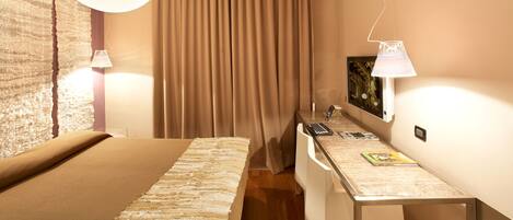 A hotel room with a bed, a desk with a lamp, a chair, a curtain, and a wall-mounted picture.