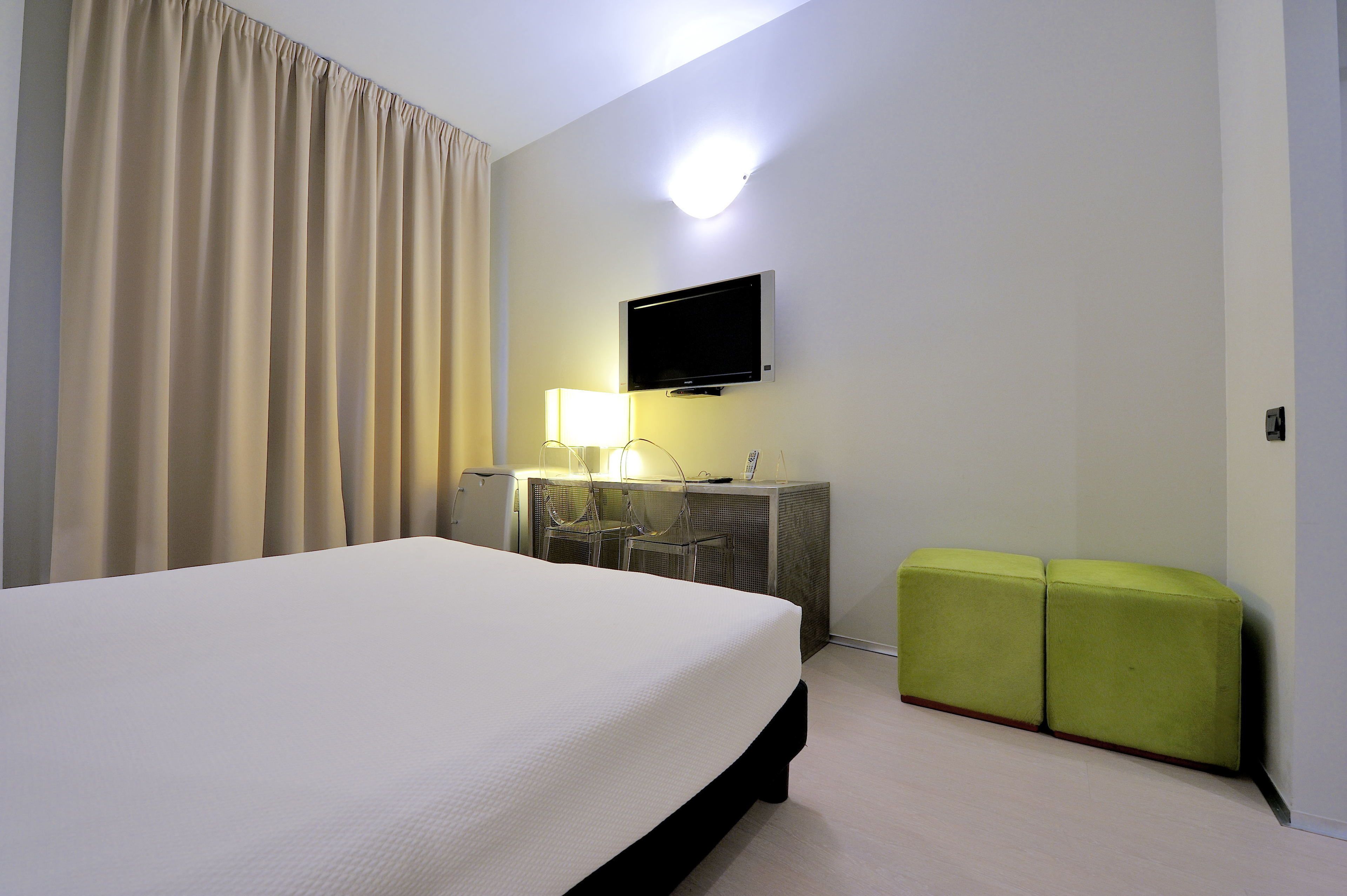 A hotel room with a bed, a desk with a lamp, a television, and two green ottomans.
