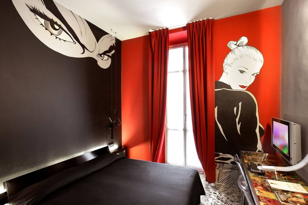 A modern hotel room with a large bed, a desk with a computer, and a window with red curtains.