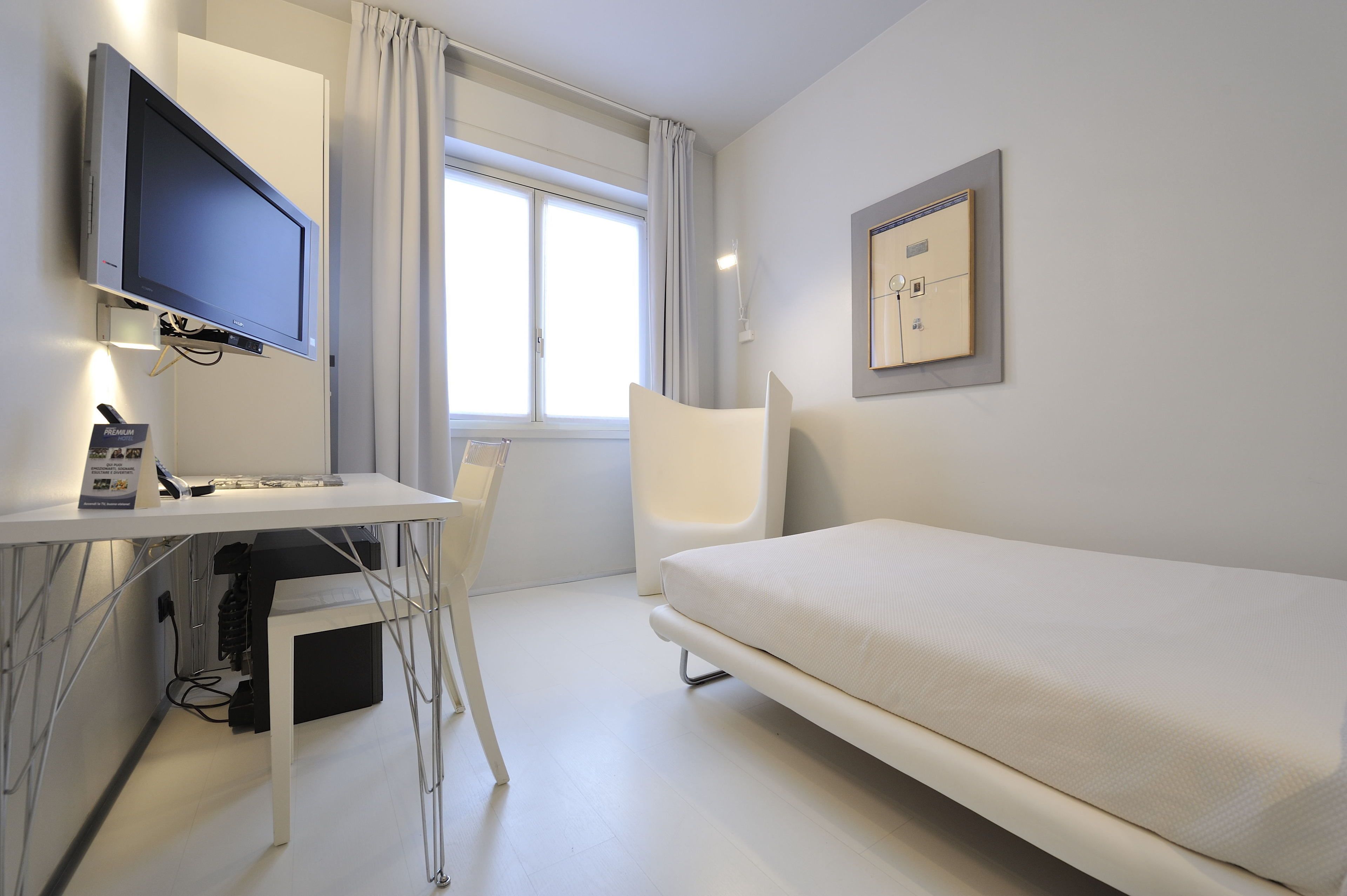 A hotel room with a bed, a desk with a computer, a chair, a TV, and a window with curtains.