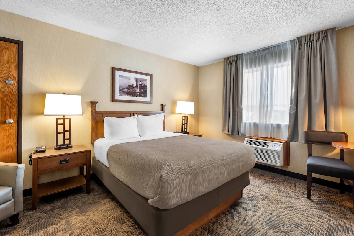 A hotel room with a large bed, two bedside tables with lamps, a chair, a desk, and a large window with curtains.