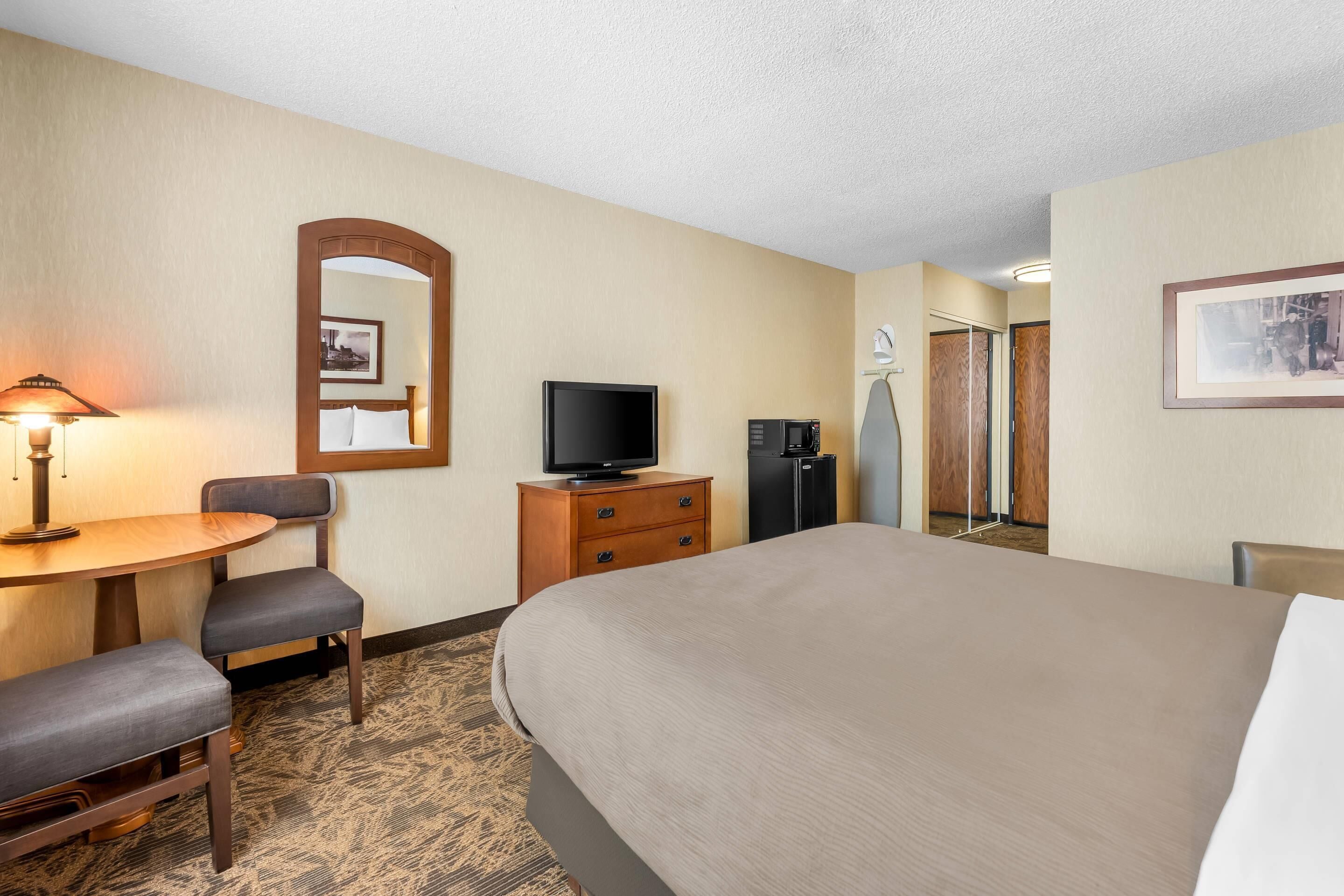 Una habitación de hotel con cama, escritorio y silla, televisor, cómoda y espejo.