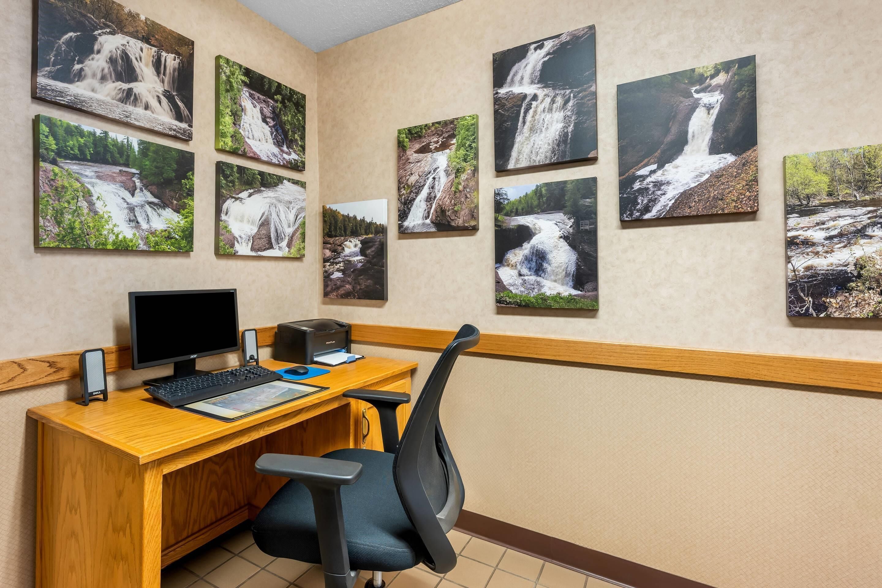 A hotel room with a wooden desk, a computer, a chair, and multiple waterfall paintings on the wall.